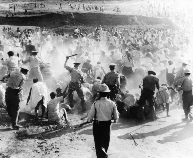 Das Bild zeigt die Massakerszene von Sharpeville. Im Vordergrund die südafrikanischen Polizisten exzessive Gewalt anwenden. Die Staubwolken in der Mitte des Bildes und die große Anzahl von Menschen, die von der Szene weglaufen. (Source from ANC archives, unknown photographer, 21st March 1960)